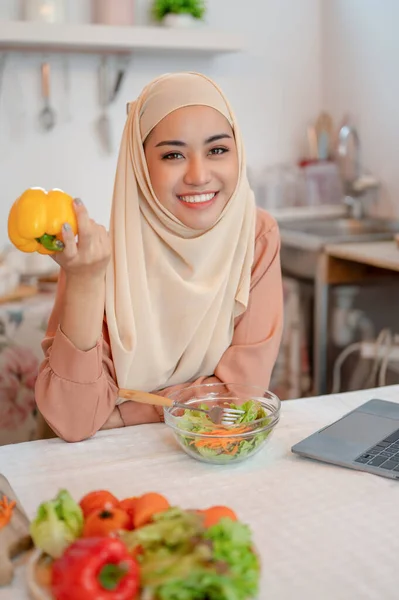 ヒジャーブの美しいアジアのムスリム女性の肖像画は 彼女の手に甘いコショウ 健康的なサラダボウル キッチンテーブルのノートパソコンとキッチンテーブルに座っています — ストック写真