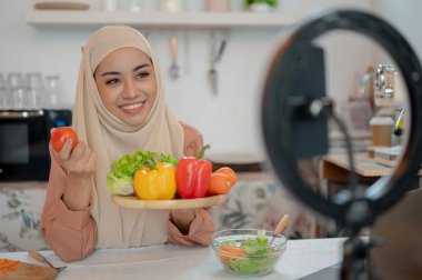 Güzel bir Asyalı Müslüman kadın yemek blogcusu ya da yaşam tarzı etkileyicisi sağlıklı yemeğinin malzemelerini kameraya gösterirken mutfakta yemek pişirme videosunu çekiyor..