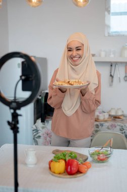 Neşeli ve güzel bir Asyalı Müslüman yaşam tarzı etkileyicisi ya da yemek blogcusu mutfakta yemek pişirme videosunu kaydederken kameraya ev yapımı waffle 'ını gösteriyor..