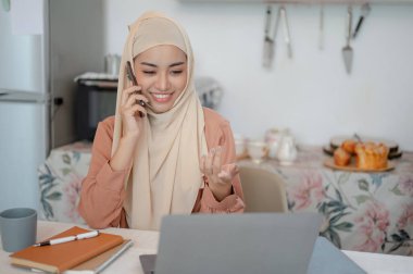 Türban ya da başörtüsü takmış güzel bir Asyalı Müslüman kadın mutfaktaki bir masada dizüstü bilgisayarıyla konuşurken telefonda konuşuyor. Evden çalışmak, uzaktan çalışmak, serbest çalışmak.