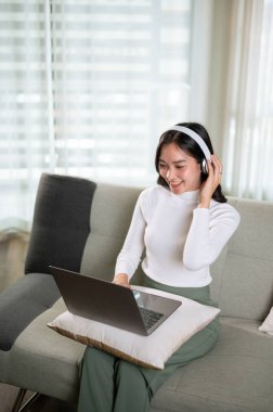 Rahat kıyafetler içindeki güzel bir Asyalı kadın oturma odasında kanepede otururken kulaklık takıyor ve dizüstü bilgisayarından bir şeyler izliyor. Video görüşmesi, film izleme, online görüşme.