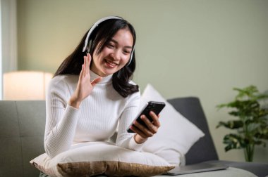 Neşeli ve güzel, kulaklık takan Asyalı bir kadın oturma odasındaki bir kanepede dinlenirken akıllı telefonuyla arkadaşlarıyla sohbet etmekten zevk alıyor..