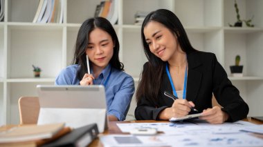 Genç ve güzel bir Asyalı kadın ofis çalışanı meslektaşı ile birlikte tablet üzerinde çalışıyor ve ofisteki mali verileri analiz ediyor..