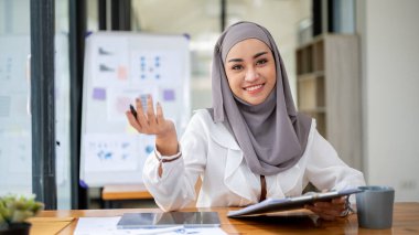 Güzel, gülümseyen, tesettürlü, tesettürlü Müslüman bir iş kadınının portresi modern bir ofiste masasında oturuyor..
