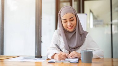 Başörtüsü ya da türban takan çekici bir Asyalı iş kadını formları dolduruyor, belgeleri imzalıyor ya da modern bir ofiste masasında kağıt üzerine notlar alıyor..