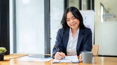 Çekici bir Asyalı genç iş kadını formları dolduruyor, belgeleri imzalıyor ya da modern bir ofisteki masasında notlar alıyor..