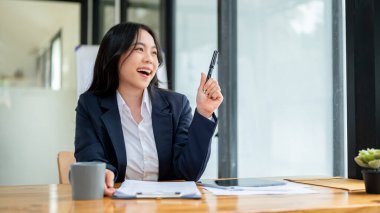 Çekici ve mutlu Asyalı genç bir iş kadını ya da kadın ofis çalışanı masasında oturur, pencereden dışarı bakar, yeni yaratıcı fikirleri düşünür.