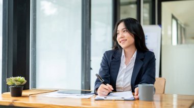 Çekici ve mutlu Asyalı genç bir iş kadını ya da kadın ofis çalışanı masasında oturur, pencereden bakar, yeni fikirleri düşünür ve kariyer başarısını hayal eder..