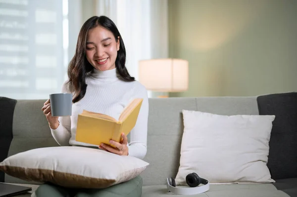 一位穿着舒适衣服的美丽而快乐的亚洲女人喜欢看书 而她早上的咖啡就在客厅的沙发上 业余爱好 家庭生活 — 图库照片