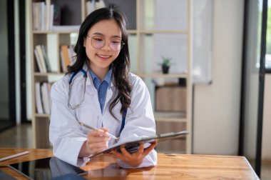 Beyaz önlüklü, yakışıklı ve profesyonel bir Asyalı kadın doktor tıbbi vaka bilgilerini bir panoda kontrol ediyor. Hastanedeki ofisinde masasında çalışıyor..