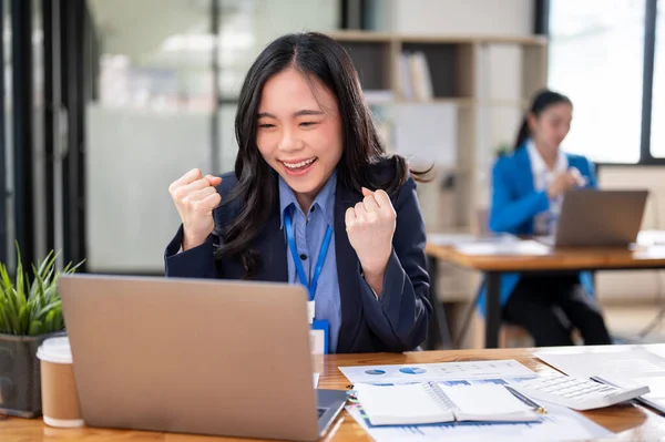 A cheerful young Asian businesswoman is looking at her laptop screen with a happy face, receiving good news online, celebrating her success, and rejoicing while sitting at her desk.