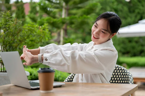 A beautiful and relaxed young Asian woman is stretching her arms while working remotely at a cafe\'s outdoor seating area. Urban lifestyle concept
