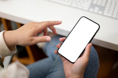 Ofisteki masasında akıllı telefonunu kullanan sıradan giyinmiş bir kadının yakın plan görüntüsü. Akıllı telefon beyaz ekran modeli. İnsanlar ve teknoloji kavramı