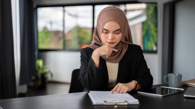 Profesyonel ve odaklanmış Asyalı Müslüman iş kadını tesettür takmış ofisinde iş belgelerini okuyor ve inceliyor..