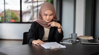 Profesyonel ve odaklanmış Asyalı Müslüman bir iş kadını tesettür takmış iş belgelerini okuyor ve inceliyor, ofisinde masasında çalışıyor..