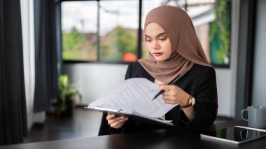 Yetişkin, Asyalı, Müslüman bir iş kadını ya da tesettürlü bir kadın yönetici, ofisindeki masasında iş belgelerini inceliyor..