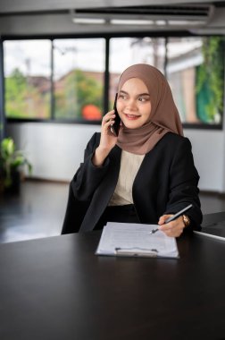 Profesyonel bir Asyalı Müslüman iş kadını ofisinde iş belgelerini incelerken telefonda konuşuyor..