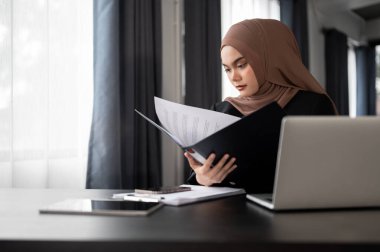 Profesyonel bir yetişkin Asyalı Müslüman iş kadını veya kadın yönetici, özel ofisindeki masasında çalışarak, bir klasördeki iş raporlarını incelemeye odaklanır..