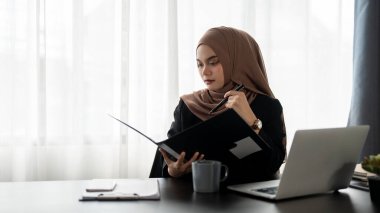 Profesyonel ve düşünceli bir Asyalı Müslüman iş kadını veya kadın yönetici masasındaki bir klasördeki iş raporlarını incelemeye odaklanır..