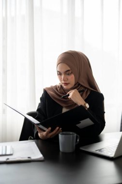 Profesyonel ve düşünceli Asyalı Müslüman bir iş kadını ya da tesettürlü kadın patron bir klasördeki raporlara odaklanıyor, ayrıntıları dikkatlice kontrol ediyor, modern ofisindeki masasında çalışıyor..