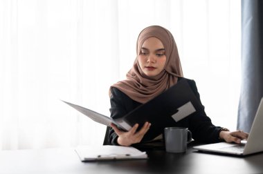 Profesyonel bir Asyalı Müslüman iş kadını ya da tesettürlü kadın patron, bir klasördeki raporlara odaklanıyor, ayrıntıları dikkatlice kontrol ediyor, modern ofisinde masasında çalışıyor..