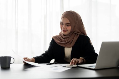 Profesyonel bir Asyalı Müslüman iş kadını ya da tesettürdeki kadın yönetici, modern bir ofiste masasındaki raporlardaki verileri inceleyerek mali raporlar üzerinde çalışıyor..