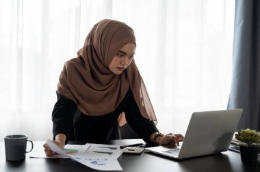 Profesyonel ve odaklanmış Asyalı Müslüman bir iş kadını finans projesi üzerinde çalışıyor, ticari mali verileri analiz ediyor ve masasında bir yatırım projesi planlıyor.