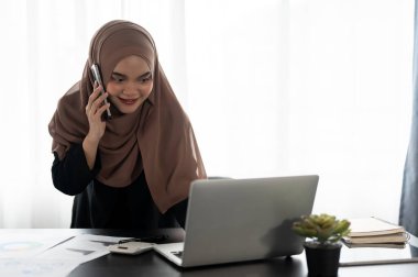 Profesyonel ve kendinden emin bir Asyalı Müslüman iş kadını ya da kadın finans asistanı, ofisinde çalışırken bilgisayarındaki detaylara bakarken müşterisiyle telefonda konuşuyor..