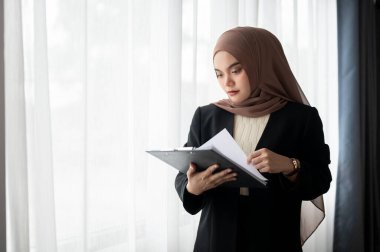 Profesyonel ve ciddi bir Asyalı Müslüman iş kadını ya da türban ya da türban takmış kadın yönetici ofisindeki pencerenin önünde dururken iş raporlarını inceliyor.