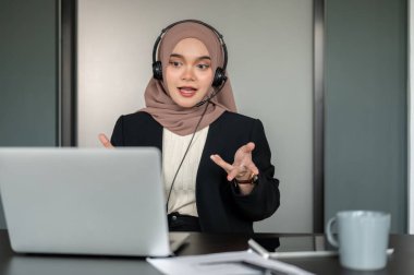 Profesyonel bir Asyalı Müslüman kadın çağrı merkezi temsilcisi veya teknik destek temsilcisi telefonla olumlu bir müşteri deneyimi sunuyor ve ofisinde çalışıyor.