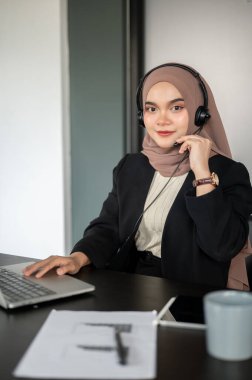 Güzel bir Asyalı Müslüman kadın satış temsilcisi ya da müşteri hizmetleri temsilcisinin portresi ofisinde otururken kameraya gülümsüyor..