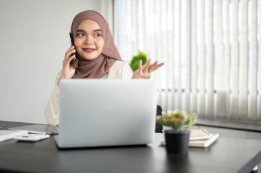 Gülümseyen ve kendinden emin Asyalı Müslüman bir iş kadını ya da tesettürlü bir kadın yönetici telefonda müşterisiyle ofisinde telefonda konuşuyor..