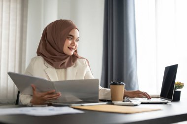 Çekici ve profesyonel Asyalı Müslüman bir iş kadını veya tesettürlü kadın yönetici bilgisayarında çalışıyor ve masasındaki bir klasörde iş raporlarını inceliyor.
