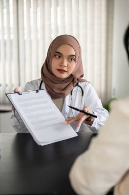 Profesyonel bir Asyalı Müslüman kadın doktorun portresi, bir hastanenin muayene odasında bir hastaya tıbbi muayene sırasında bir panoda bilgi veriyor..