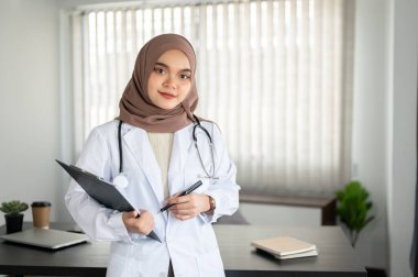Üniformalı ve tesettürlü çekici ve profesyonel bir Asyalı Müslüman kadın doktorun portresi elinde hastane veya klinikteki bir ofiste elinde bir pano ve kalemle duruyor..
