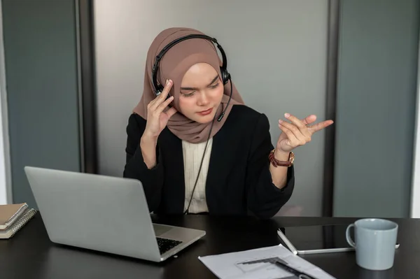 A confused and thoughtful Asian Muslim female customer call centre operator wearing a headset and hijab resolves customer issues over the call, consulting with a client over the phone.
