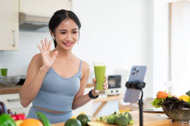 Güzel ve neşeli bir Asyalı kadın etkileyicisi veya çevrimiçi beslenme koçu canlı yayında sabah içeceğini hazırlarken ve sağlıklı yeşil smoothie tarifini online takipçileriyle paylaşırken.