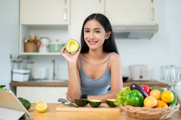 ジム服に魅力的で笑顔のアジア女性の肖像画は キッチンで彼女の健康的な食事を準備しながら カメラにアボカドを示しています — ストック写真