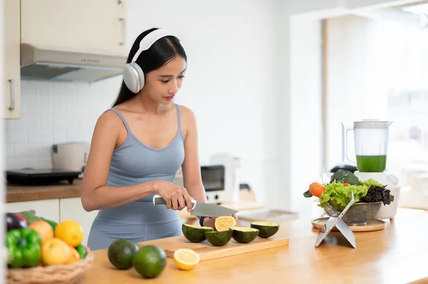 A healthy and beautiful young Asian woman in gym clothes is listening to music through her headphones while preparing her healthy breakfast after the gym in the kitchen. Urban healthy lifestyle