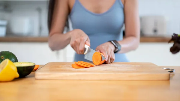Close-up image of a healthy and fit woman in gym clothes is slicing carrots on a chopping board, preparing ingredients for her healthy breakfast after the gym in the kitchen of her apartment.
