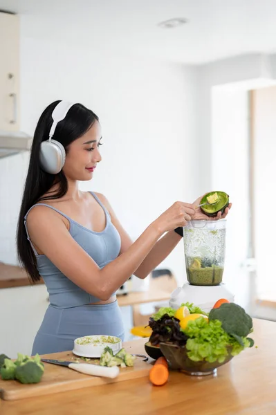 A gorgeous and slim Asian woman in gym clothes and headphones is adding an avocado to a blender, making her healthy green detox smoothie juice in the kitchen after her morning workout. Diet life