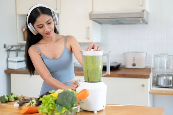 A fit and beautiful Asian woman in sportswear and headphones is making her healthy green smoothie with a blender in the kitchen. Diet, wellbeing, healthy food, urban lifestyle
