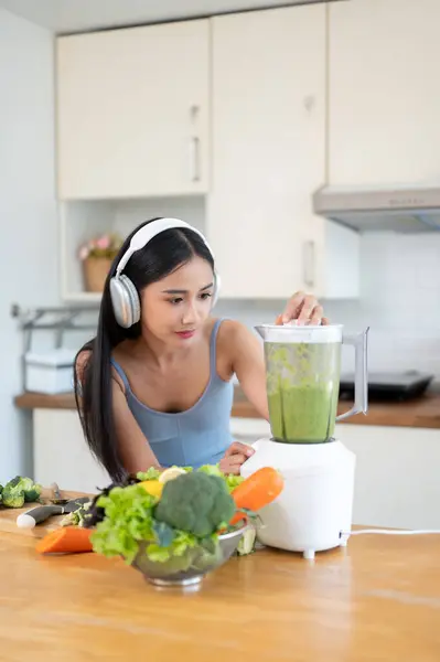 スポーツウェアとヘッドフォンの豪華でスリムなアジアの女性は 朝のトレーニングの後 キッチンのテーブルで健康な緑色のデトックススムージーを作っています ダイエット 健康的なライフスタイル — ストック写真