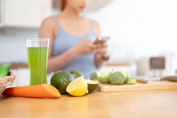 健康な緑のジュースと新鮮な果物と野菜のグラスのクローズアップ画像 キッチンの木製キッチンテーブルに女性が後ろのスマートフォンを使用して 選択フォーカス画像 — ストック写真