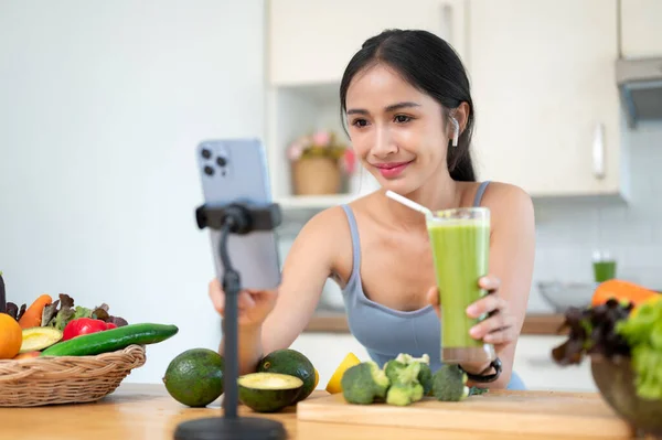 ジム服の美しく幸せなアジアの女性インフルエンサーは キッチンでそれを作っている間 彼女の健康的な緑の滑らかなレシピを共有するために彼女のソーシャルメディア上でライブストリーミングされています ダイエット グリーンジュース — ストック写真
