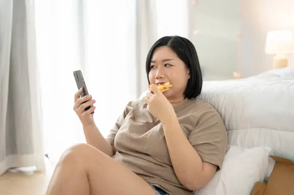 A happy and joyful Asian curvy plus-size woman in comfy clothes is enjoying pizza while scrolling through social media on her smartphone in her bedroom. Unhealthy lifestyle, domestic life, junk food