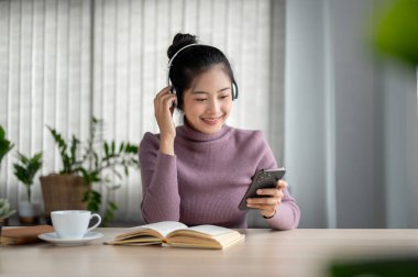 Güzel ve mutlu bir Asyalı kadın akıllı telefonuyla müzik listesi seçiyor ve odasında otururken kulaklıkla müzik dinlemekten zevk alıyor..