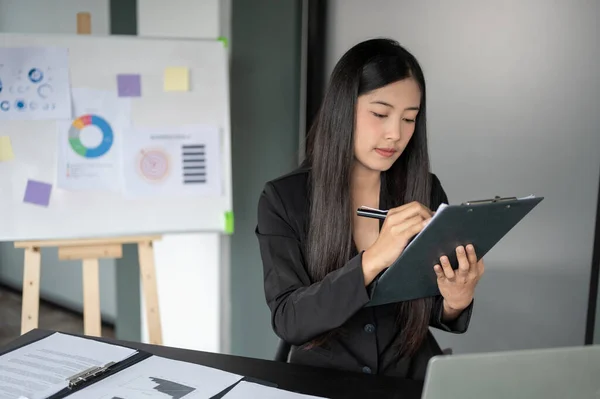 A professional millennial Asian businesswoman in a formal black suit focuses on examining business reports while working at her desk in the office.