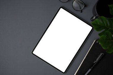Beyaz ekran dijital tablet, gözlük, kahve fincanı ve koyu gri çalışma alanı arka planında kırtasiye malzemesi. Üst görünüm, düz uzanma