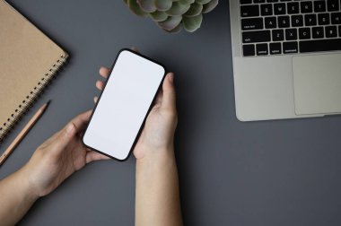 Beyaz ekran akıllı telefonu elinde tutan bir kadının üst görüntüsü modern koyu gri ofis masasının üzerinde bir dizüstü bilgisayar, bir kalem ve bir defter ile. Çalışma alanı, düz yatak